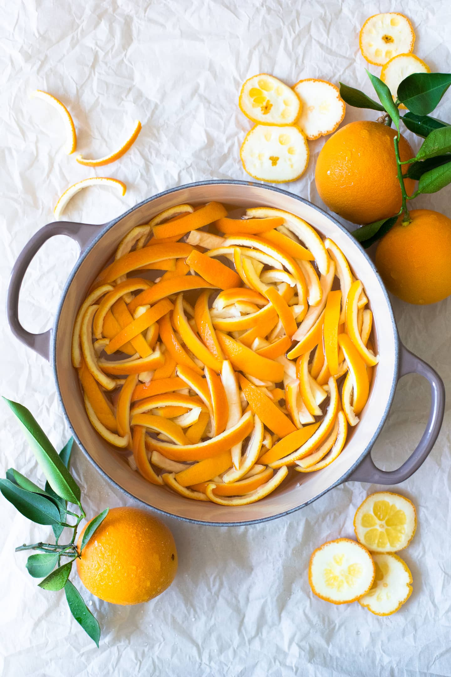 Overhead view of strips of orange peel covered with water in a pot, surrounded by more oranges and peel cuttings.