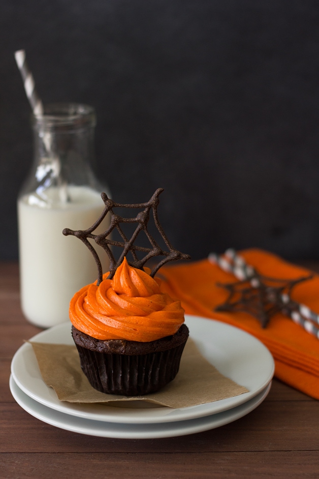 Front-facing view of one Pumpkin Halloween Cupcake featuring pumpkin-chocolate cake with orange-colored cream cheese frosting topped with a chocolate spiderweb, sitting on top of two stacked white plates. A bottle of milk  and grey paper straws are in the background.