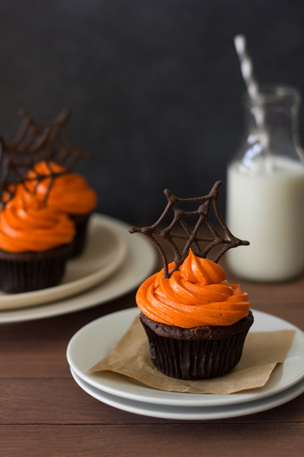 Front-facing view of a Pumpkin Halloween Cupcake featuring pumpkin-chocolate cake with orange-colored cream cheese frosting topped with a chocolate spiderwebs. Two more cupcakes and a bottle of milk are in the background.