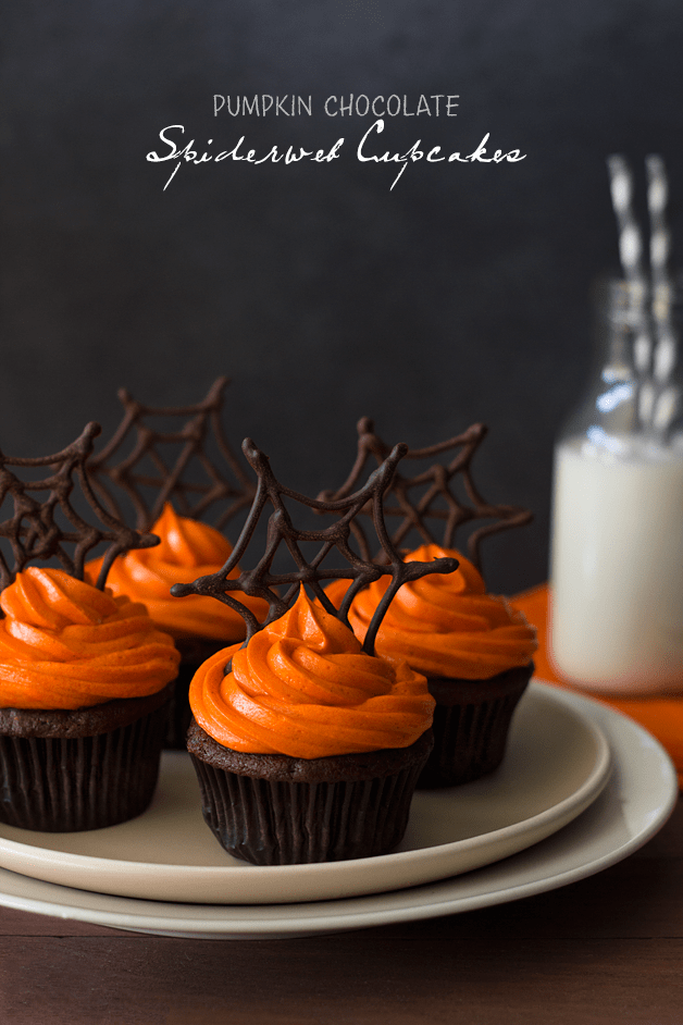 Front-facing view of pumpkin Halloween cupcakes featuring pumpkin chocolate cupcakes with orange-colored cream cheese frosting and topped with chocolate spiderwebs, plus a glass of milk in the background.