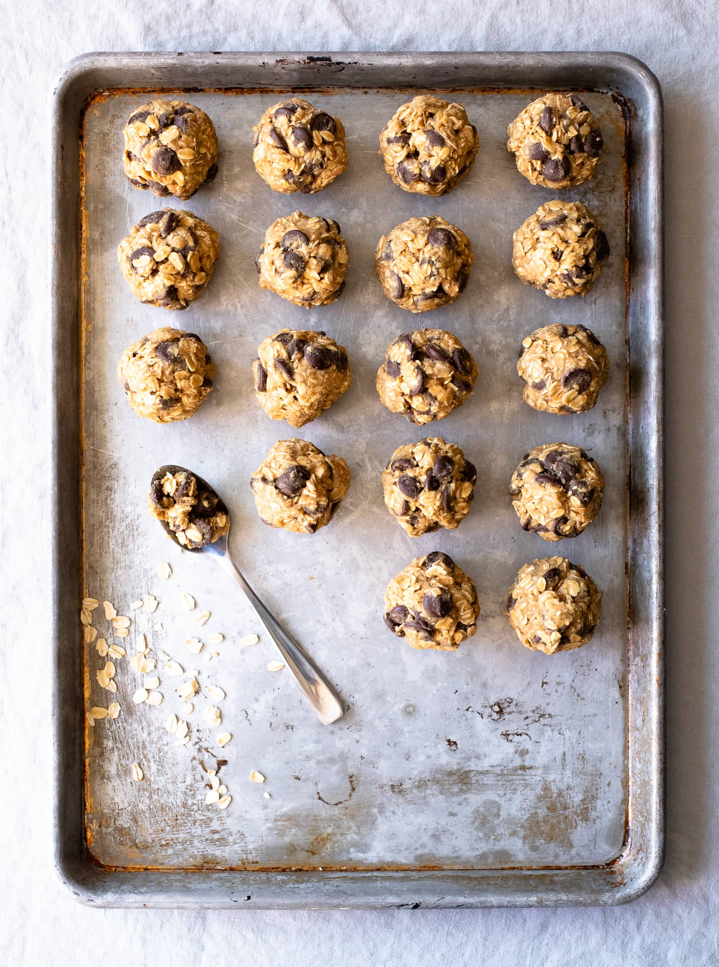 Overhead view of gluten-free oatmeal chocolate chip cookie dough balls, arranged in rows on an old metal cookie sheet, along with a spoon used for scooping the dough.