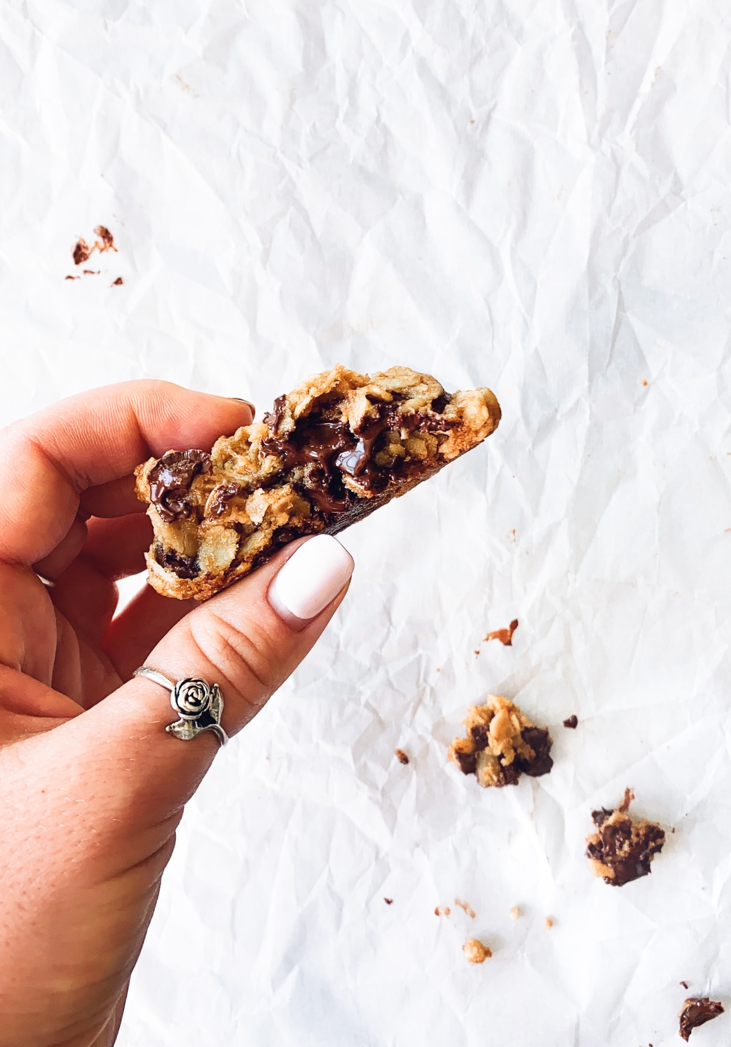 Female hand holding half of a gluten-free Oatmeal Chocolate Chip Cookie over a crinkled sheet of white parchment paper, with cookie crumbs below.
