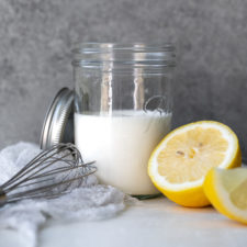 Homemade buttermilk in a mason jar, alongside slices of lemon and a whisk, against a cement backdrop.