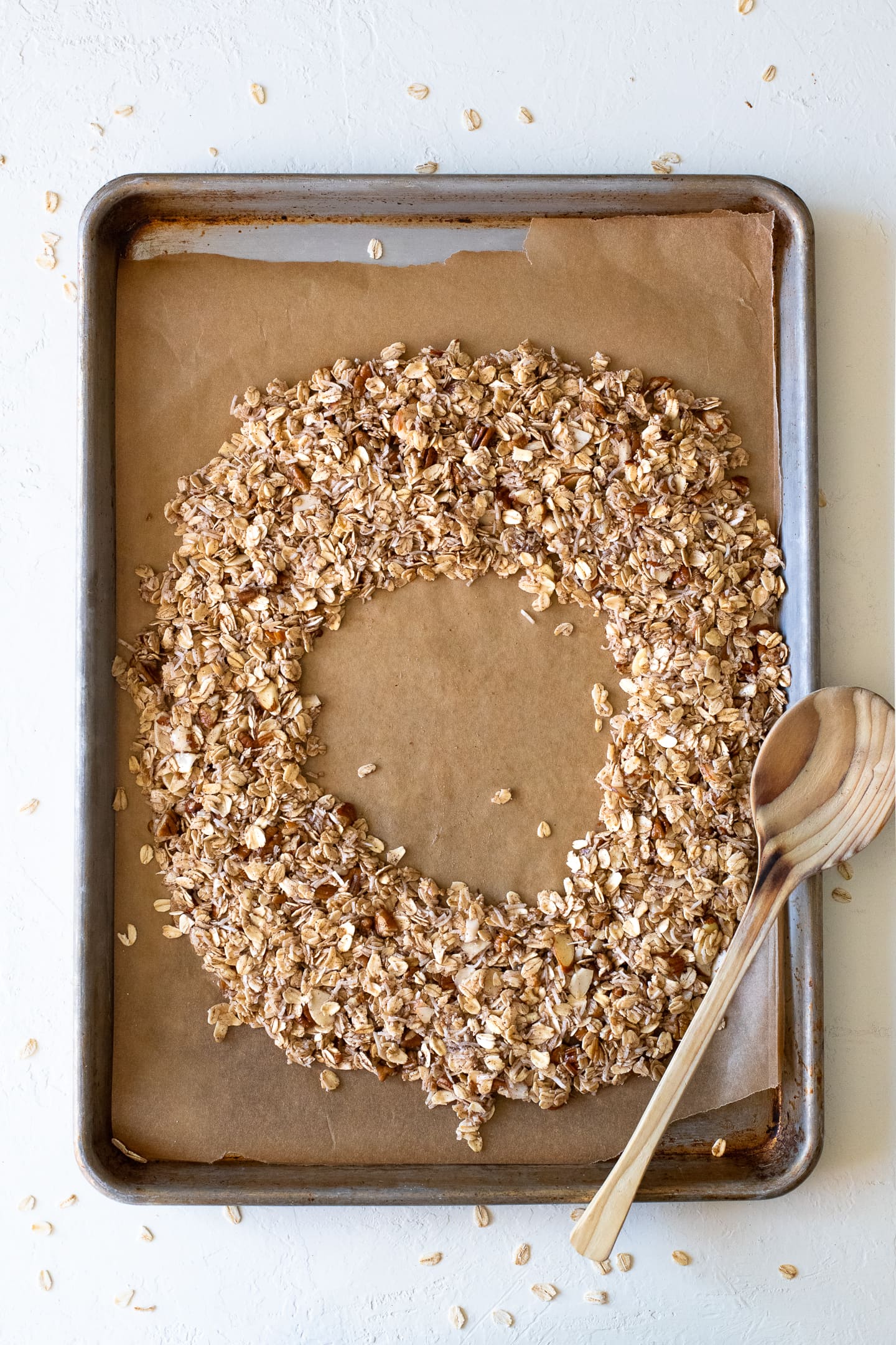 Overhead view of unbaked granola arranged in a circular shape on a sheet on a baking sheet, with a wooden spoon laying across the top.