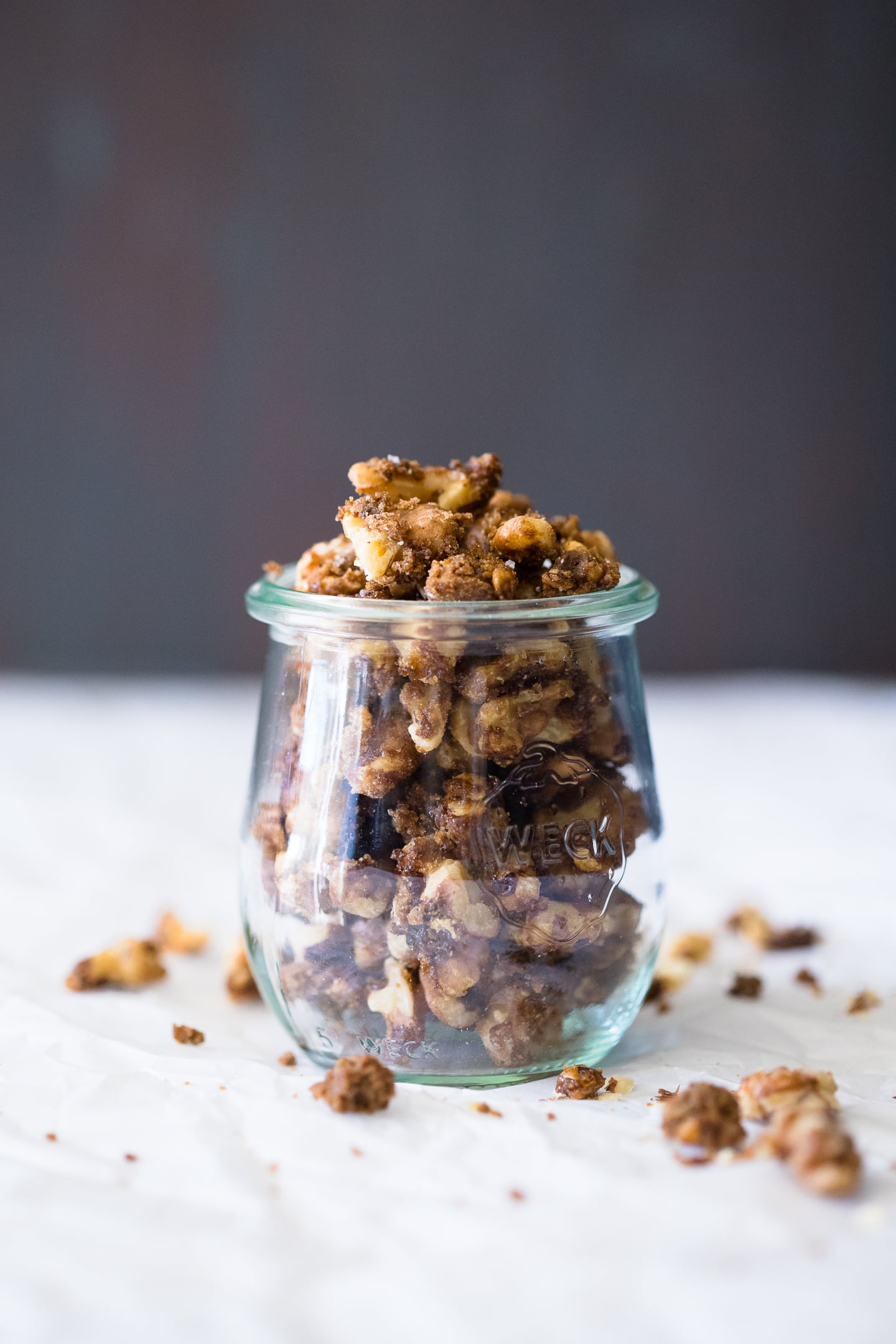 Front-facing close-up of candied walnuts in a Weck Jar, sitting on a piece of parchment paper with scattered walnut pieces surrounding it.