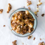 Overhead view of brown sugar candied walnuts in a jar, with more walnuts scattered on a sheet of parchment paper below.