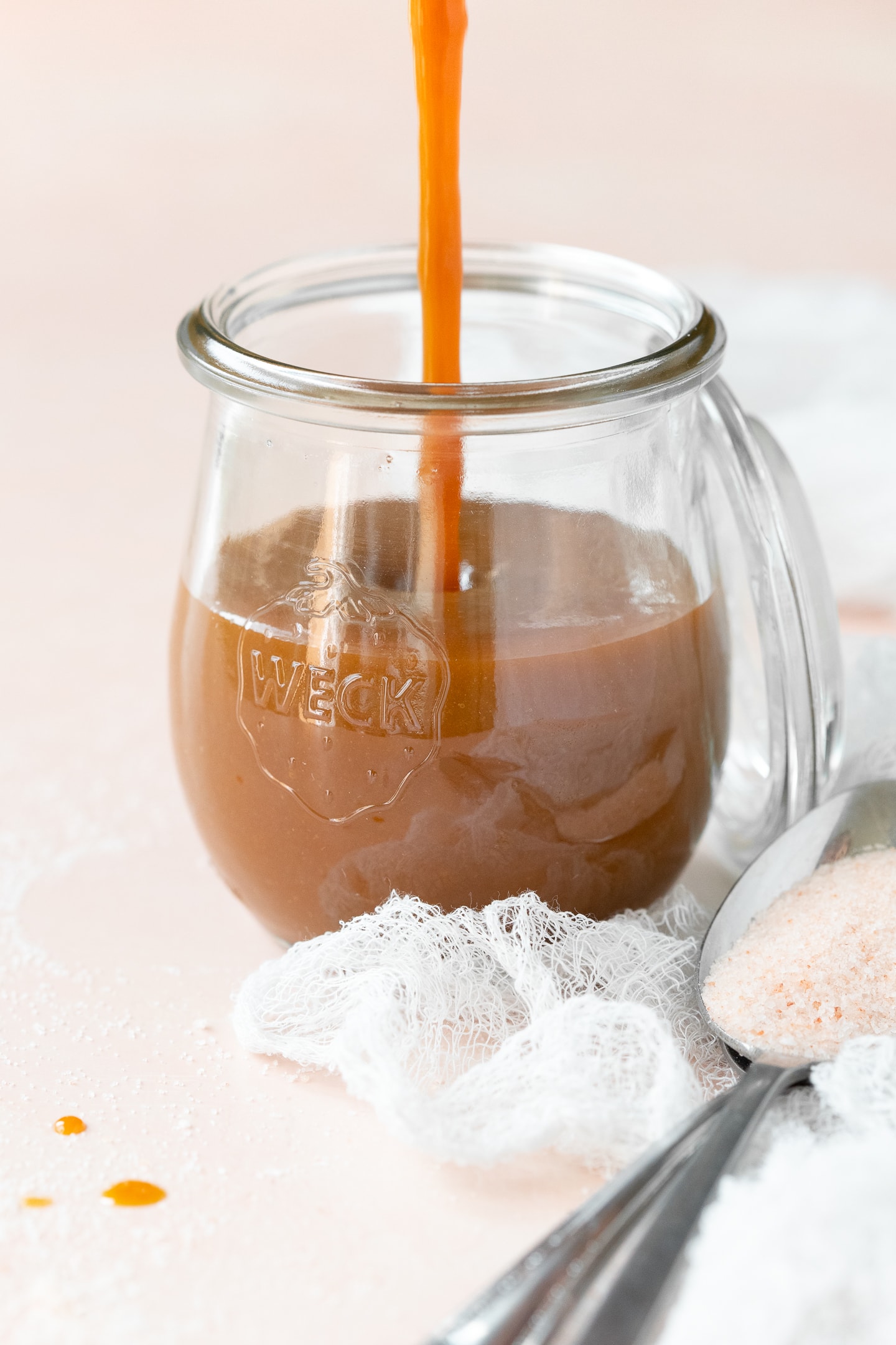 Front-facing view of caramel sauce being poured into a jar against a light pink backdrop.