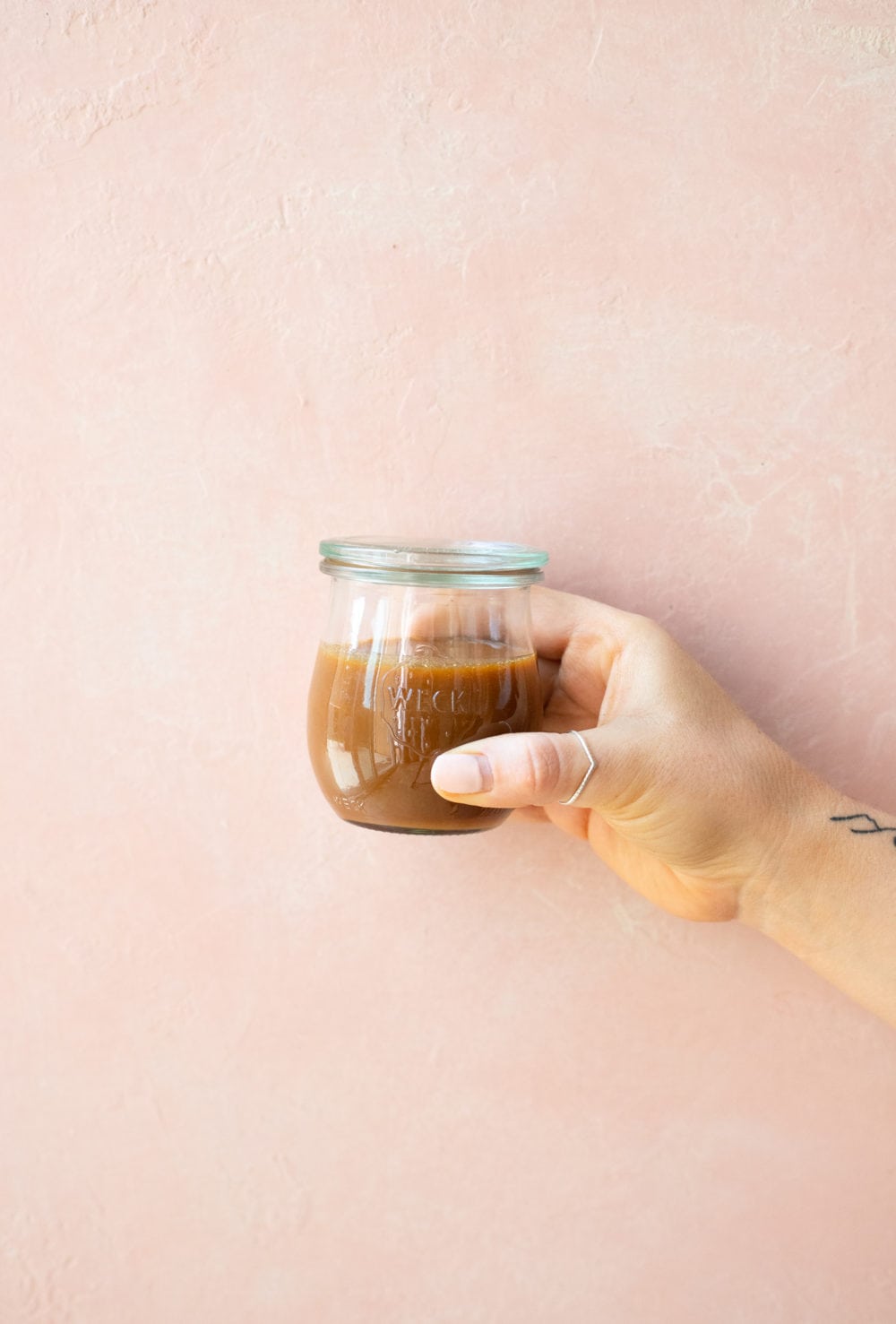 Front view of a hand holding a jar of caramel sauce against a light pink background.