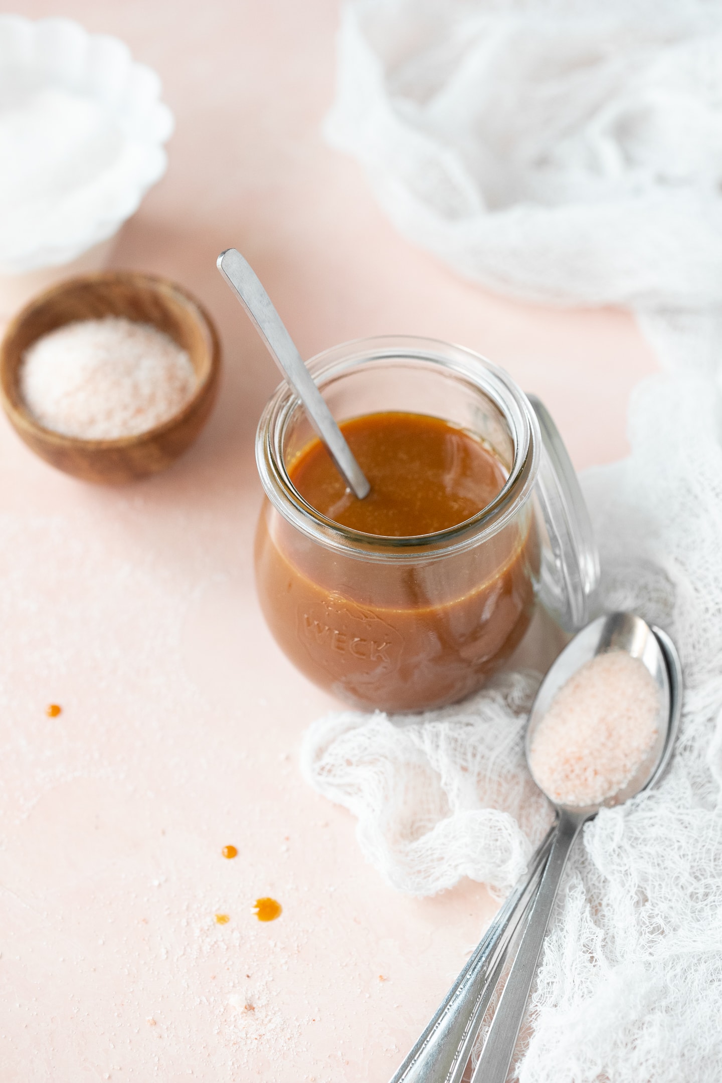 Front angled view of a jar of caramel sauce alongside bowls of sugar and sea salt, with a spoon of sea salt nearby.