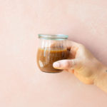Front view of a hand holding a jar of caramel sauce against a light pink background.
