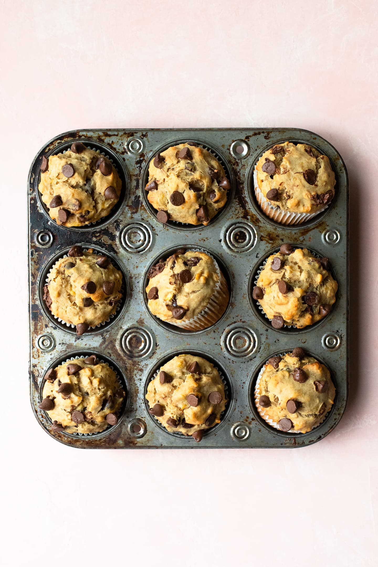 Overhead view of Peanut Butter Banana Chocolate Chip Muffins in a muffin pan on a light pink backdrop.