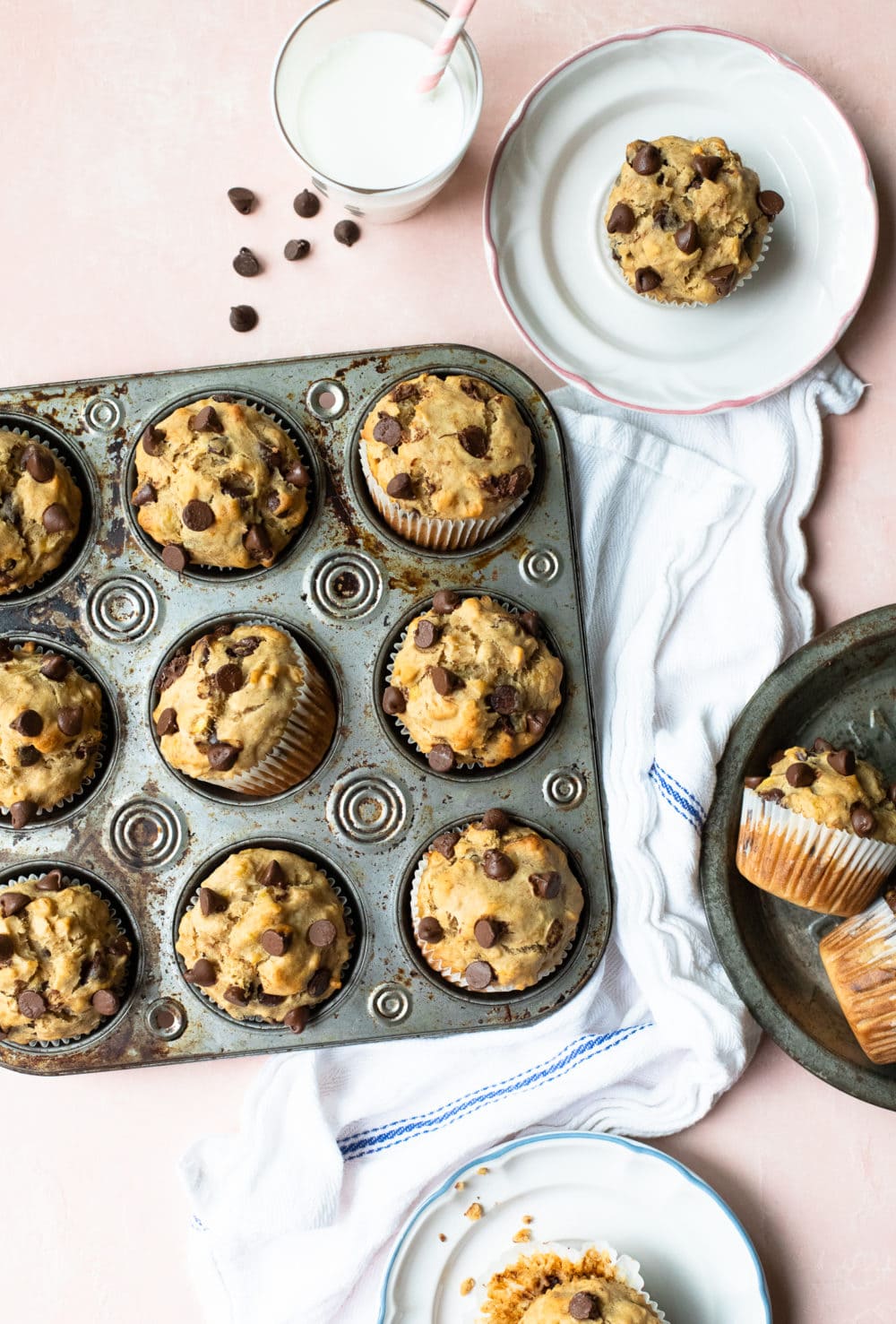 Overhead view of Peanut Butter Banana Chocolate Chip Muffins in a muffin tray, along with extra muffins on plates, over a light pink backdrop.