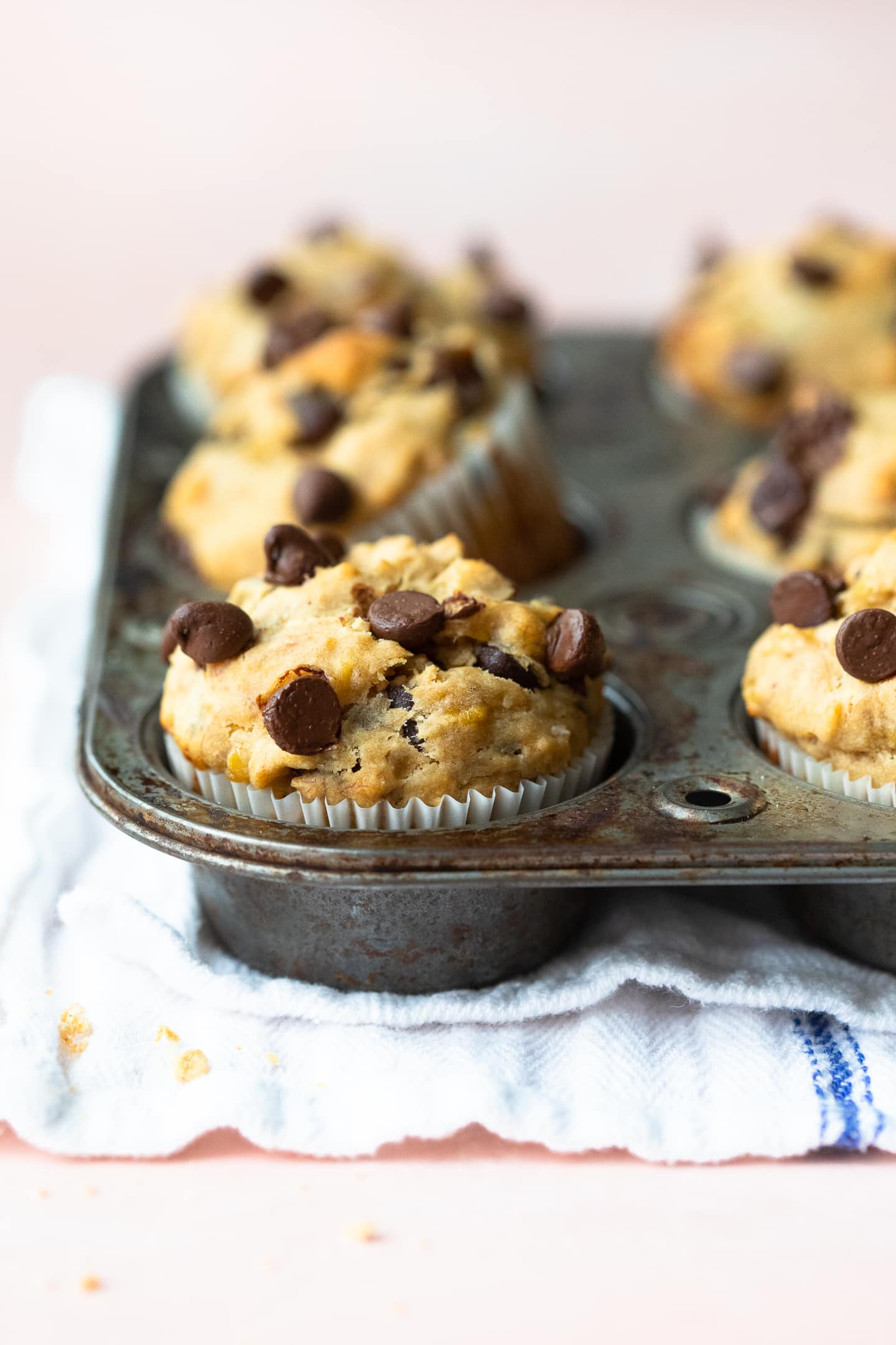 Front-facing view of Peanut Butter Banana Chocolate Chip Muffins in a muffin pan, with a white linen underneath, on a light pink backdrop.