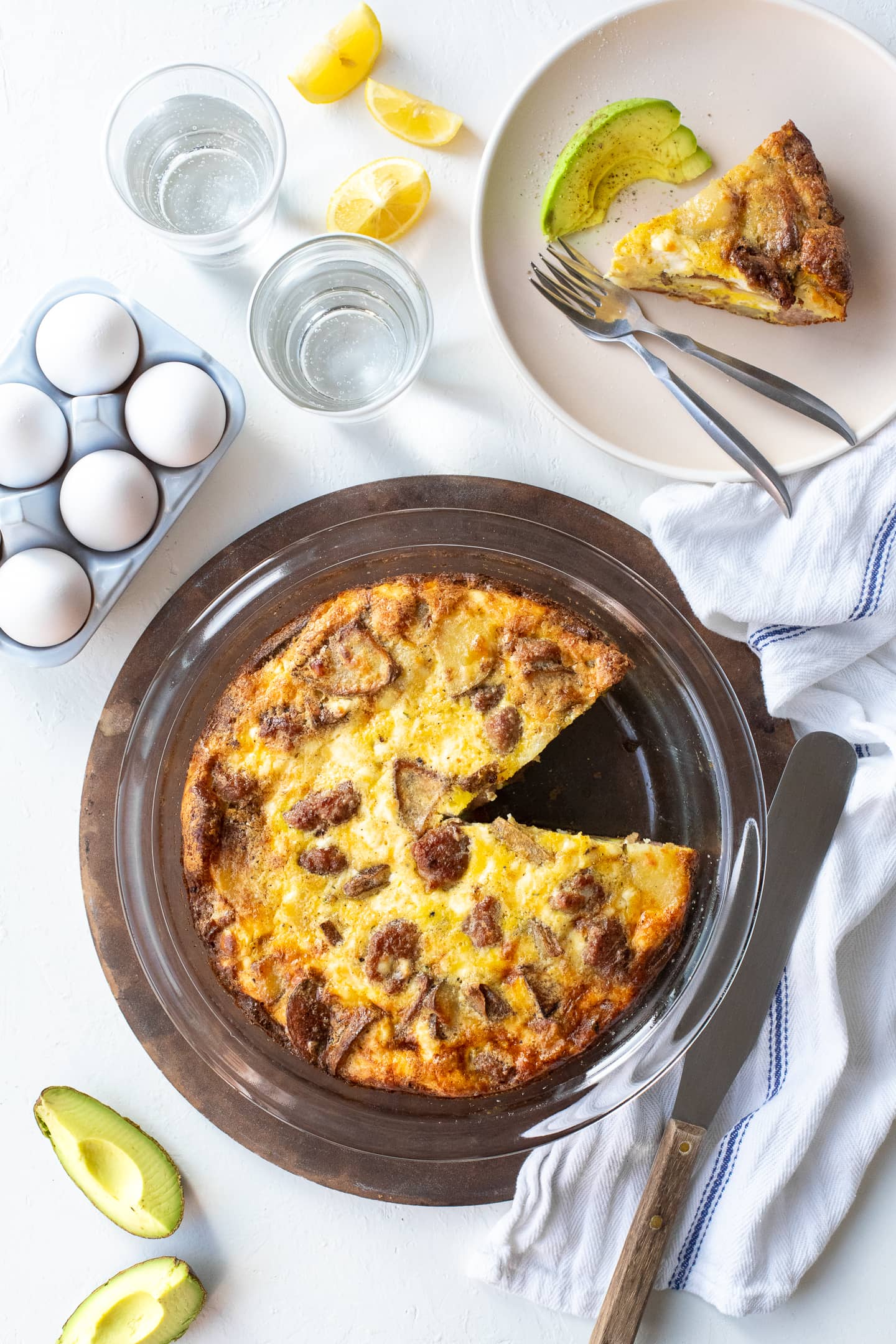 Overhead view of crustless quiche in a pie dish with a slice removed, surrounded by eggs, avocado, and a slice ready to serve on a plate.