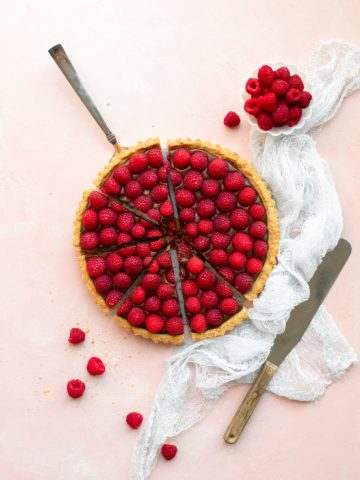 Overhead view of a Raspberry Chocolate Tart, sliced and ready to serve, surrounded by extra raspberries, a white linen, and a pallet knife.