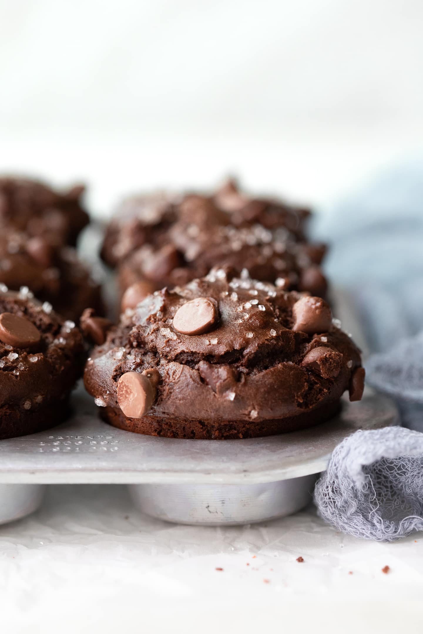 Close-up view of double chocolate muffins in a muffin pan, alongside a grey linen over a white backdrop.