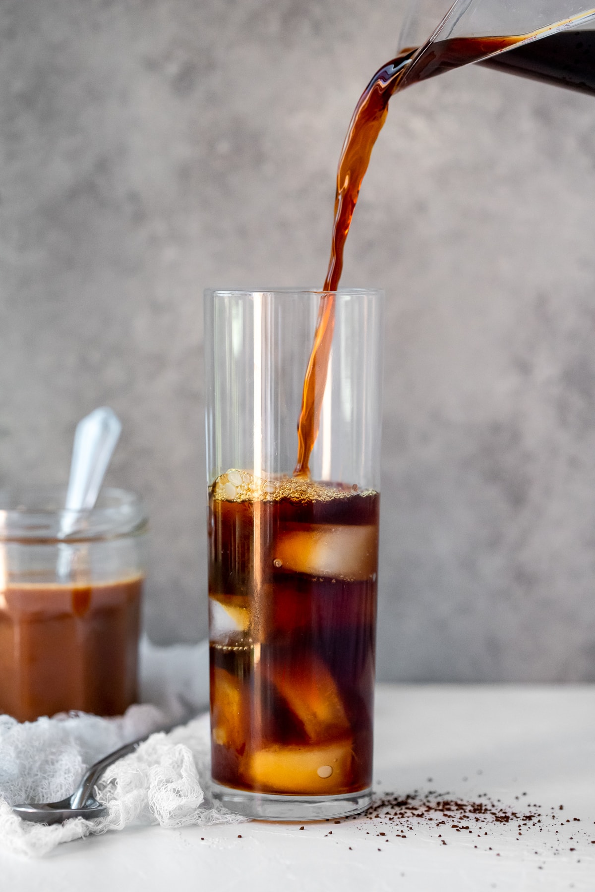 Café a ser vertido sobre gelo num copo de vidro alto, com um frasco de caramelo no fundo.