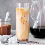 Caramel iced coffee in a tall glass, with a jar of caramel and a pour over full of coffee in the background.
