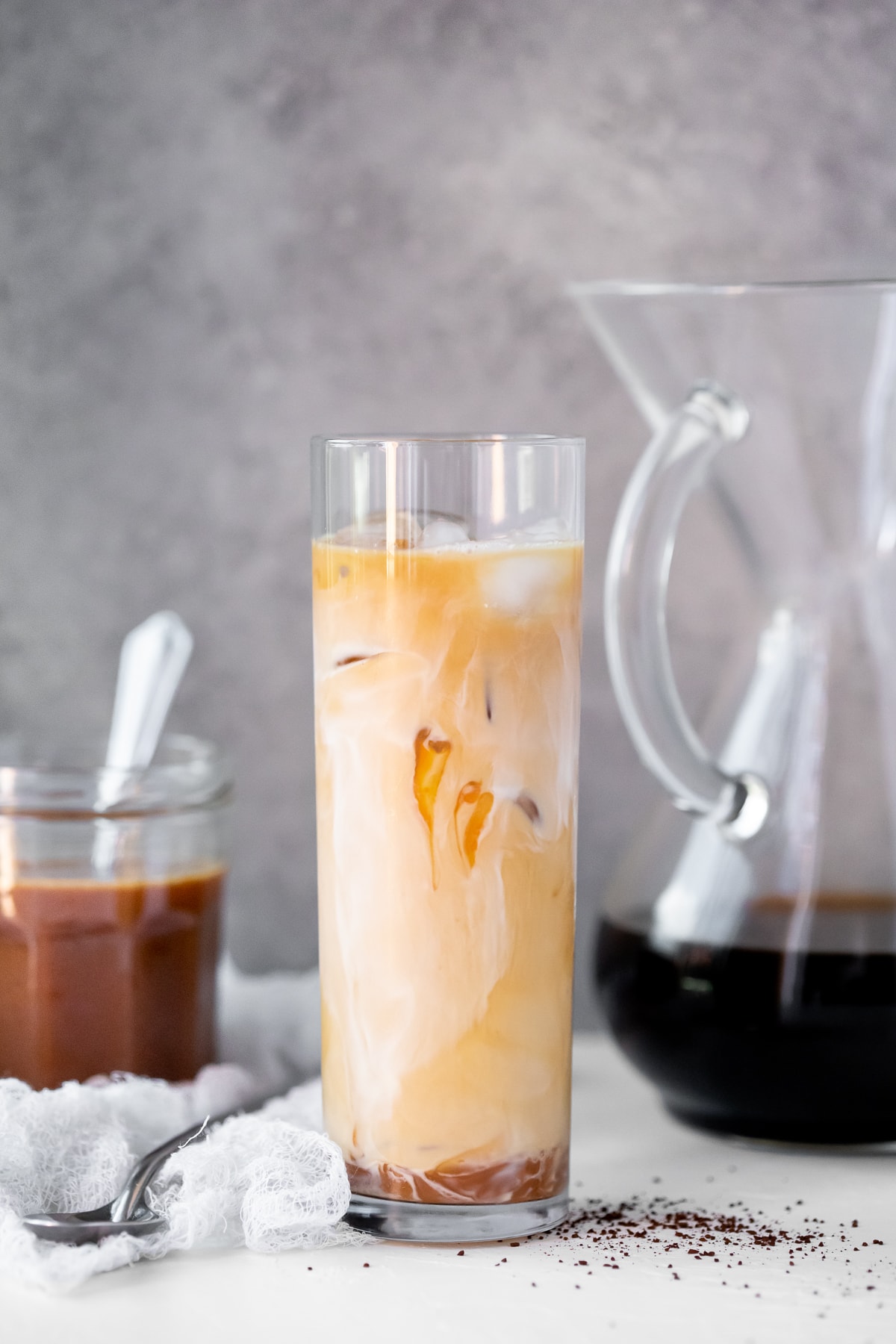Caramel iced coffee in a tall glass, with a jar of caramel and a pour over full of coffee in the background.