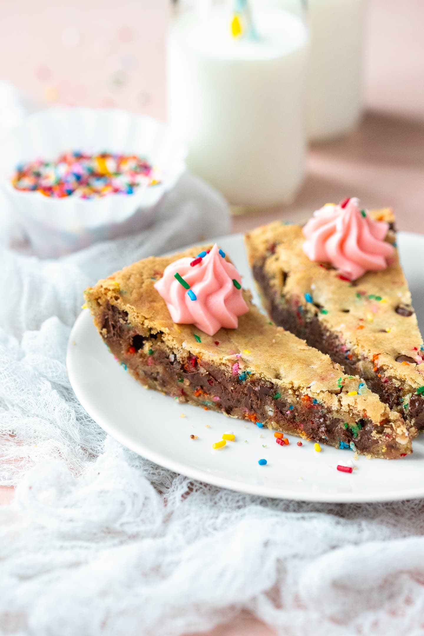 Front-angled view of two slices of cookie cake on a plate, with glasses of milk and a bowl of sprinkles in the background.