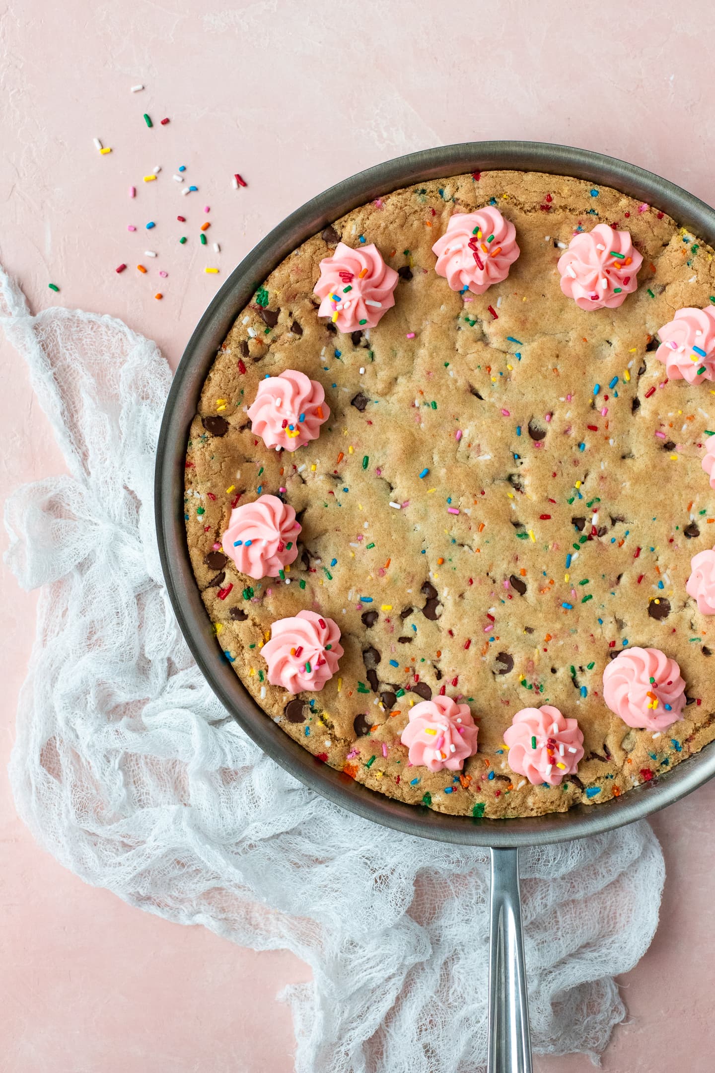 Overhead view of a chocolate chip cookie cake with a white linen to the side, over a light pink backdrop.