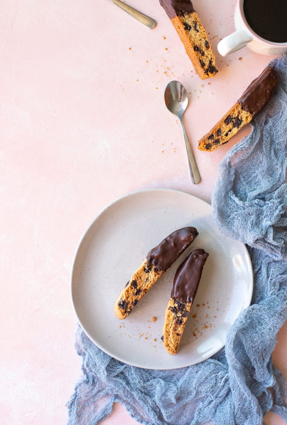 Two chocolate chip biscotti on a grey plate, surrounded by a blue-grey linen, scattered biscotti, and a cup of coffee.