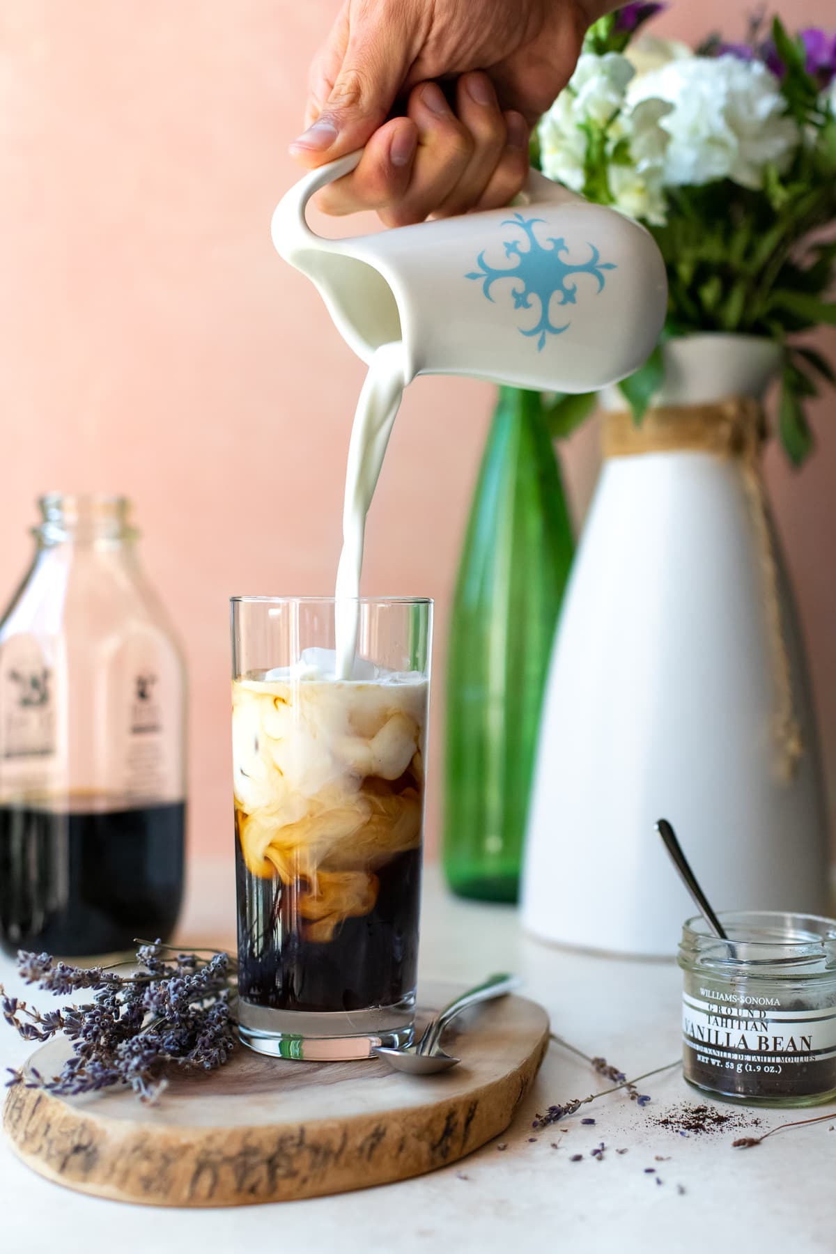 Cream being poured into iced coffee, with fresh flowers in the background against a pink backdrop.