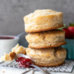 Front-facing view of 3 biscuits stacked atop each other, with a spoonful of jam in for foreground and a basket of strawberries in the background.