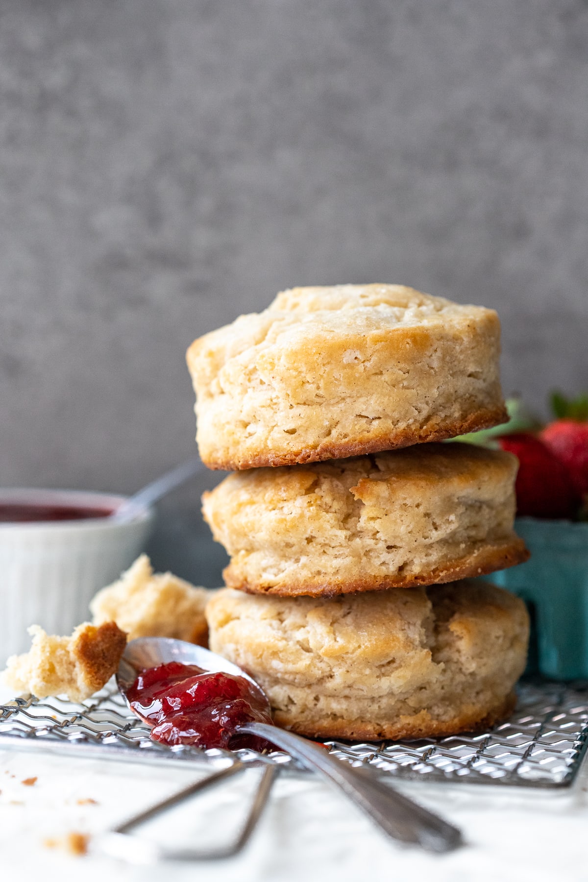 Front-facing view of 3 biscuits stacked atop each other, with a spoonful of jam in for foreground and a basket of strawberries in the background.