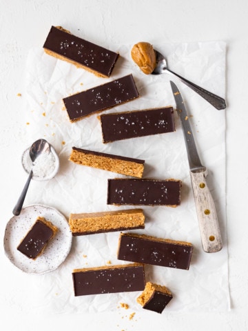 Overhead view of protein bars scattered on a sheet of parchment paper, alongside a knife and a small bowl of sea salt.