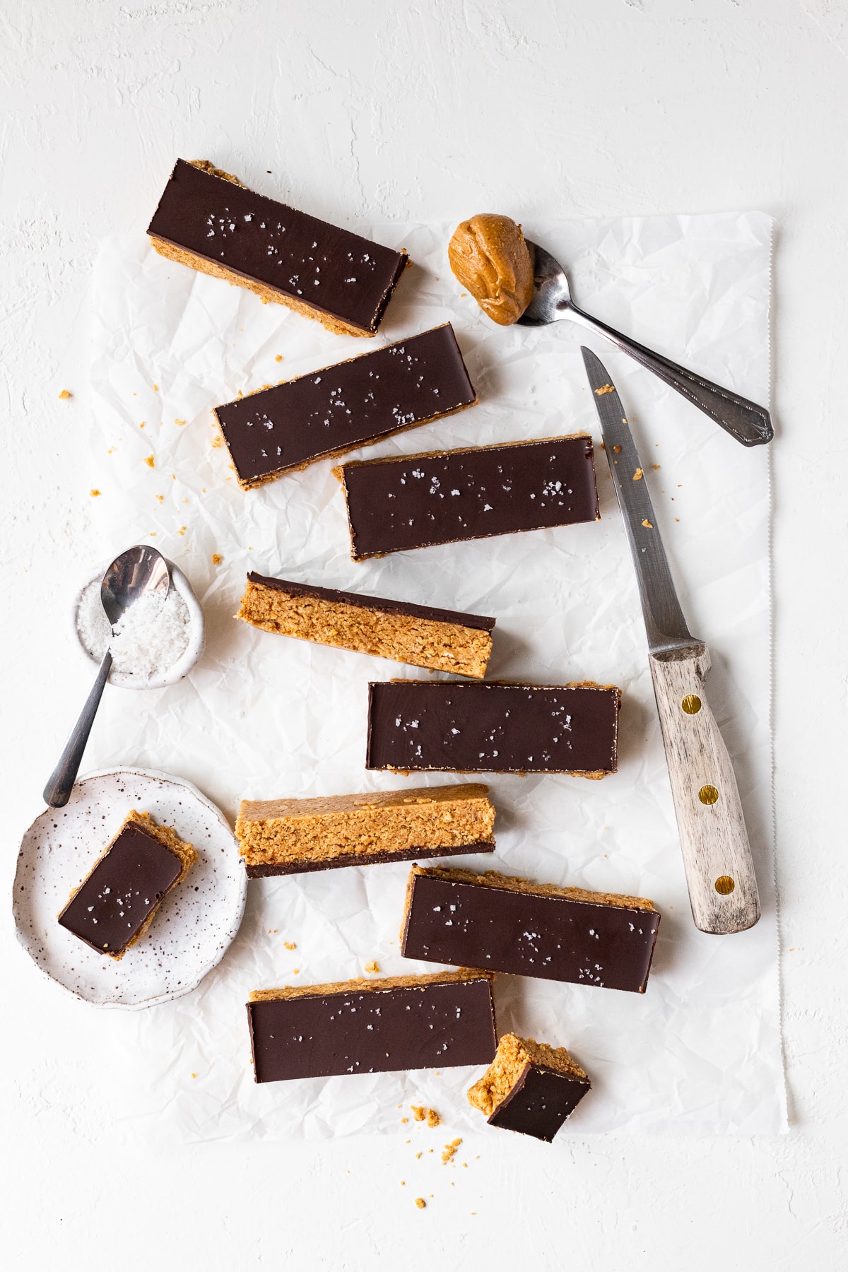 Overhead view of chocolate peanut butter protein bars scattered on a sheet of parchment paper, alongside a knife and a small bowl of sea salt.