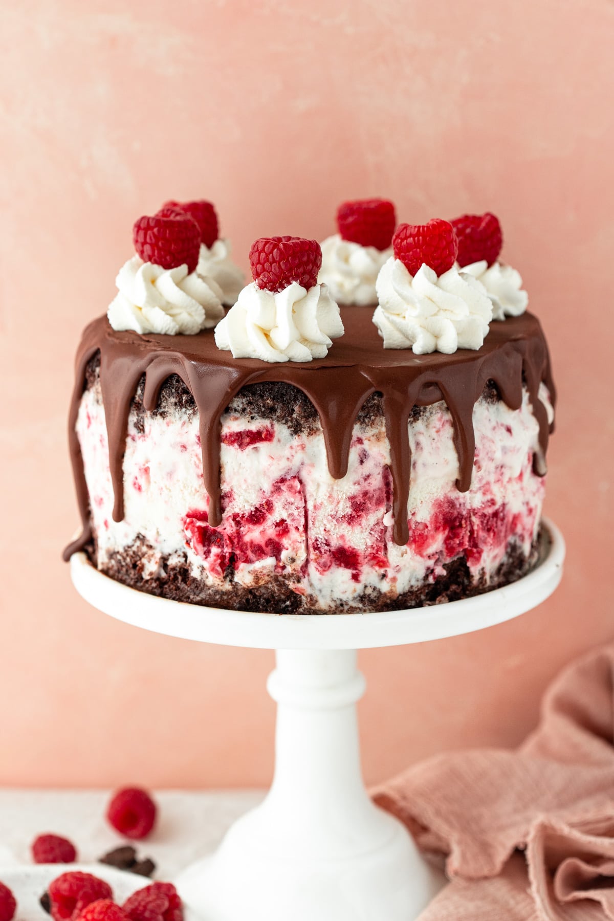 Ice cream cake on a cake stand against a pink backdrop, with raspberries in the foreground.