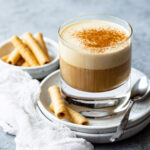 Coffee in a glass topped with cinnamon, served with rolled cookies.