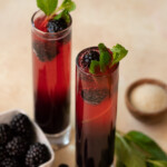 Two mimosa in champagne flutes garnished with fresh blackberries and mint.