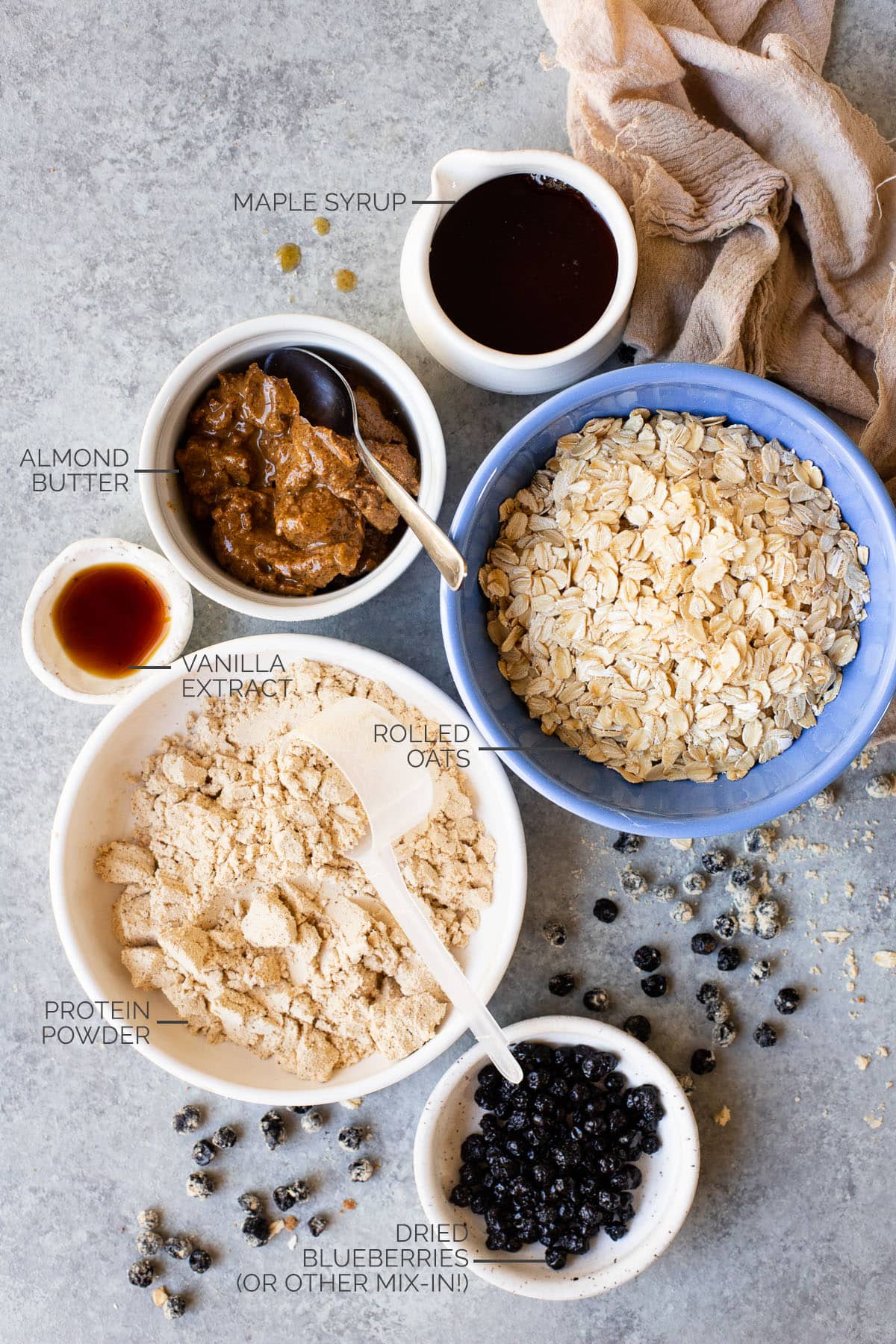 Maple syrup, almond butter, oats, protein powder, vanilla, and dried blueberries in ingredient bowls.