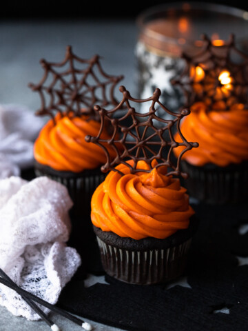 Three cupcakes with orange frosting topped with chocolate webs.