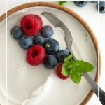 Close up of dairy-free whipped cream in a bowl with fresh berries and mint. Text reads "Dairy-Free Whipped Cream".
