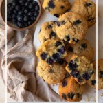 Blueberry muffins piled on a white marble cutting board, with one cut in half. Image has text overlay of recipe title.