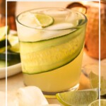 Close-up of cocktail in a rocks glass, garnished with a cucumber ribbon and lime wedge. Text overlay reads "Cucumber Moscow Mule".