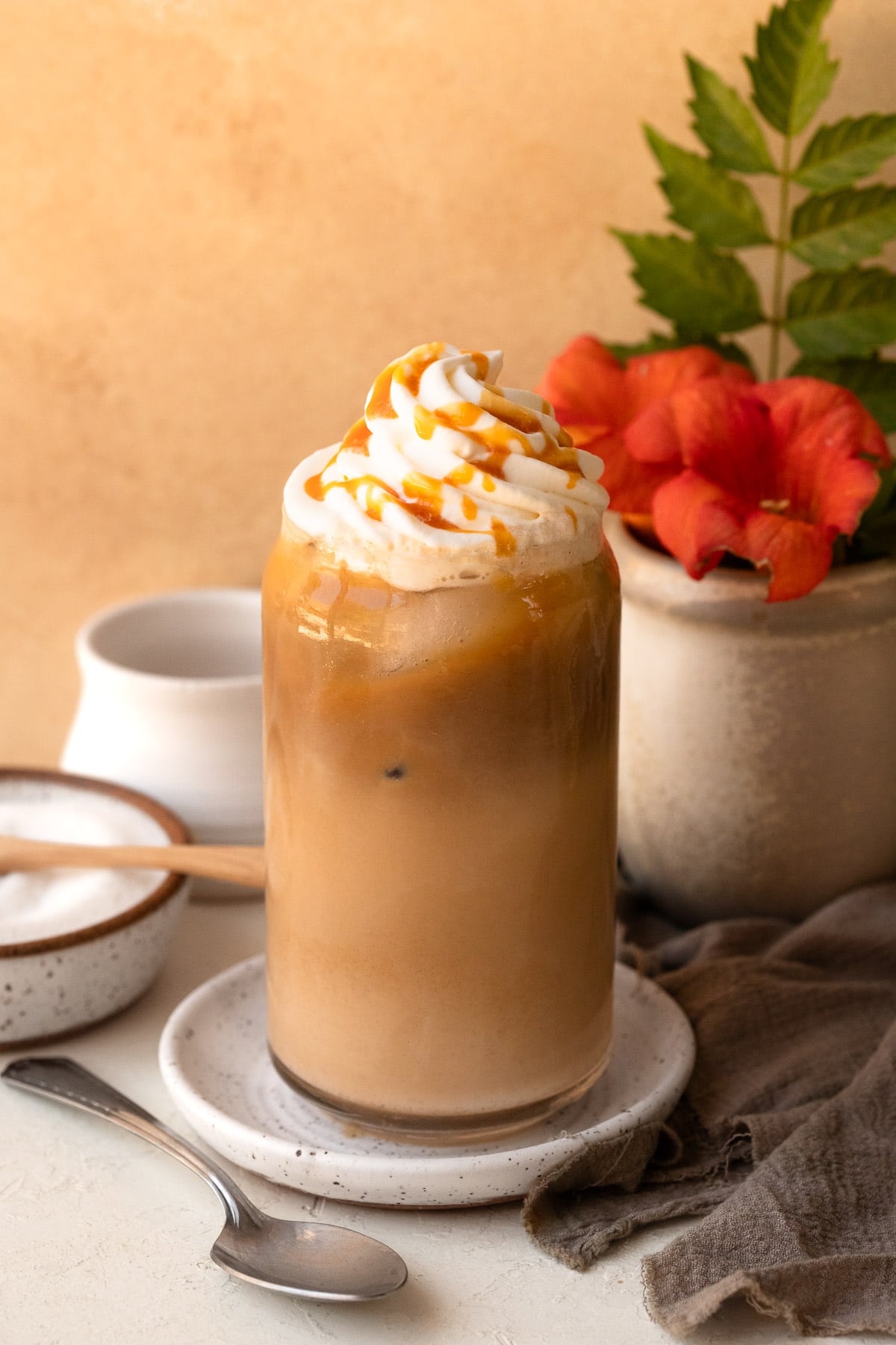Iced latte topped with whipped cream and a caramel drizzle, with a vase of flowers in the background.