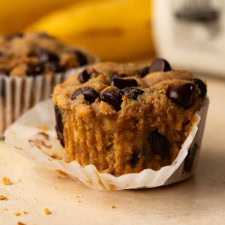 Close up of a muffin with the paper liner partially removed and bananas in the background.