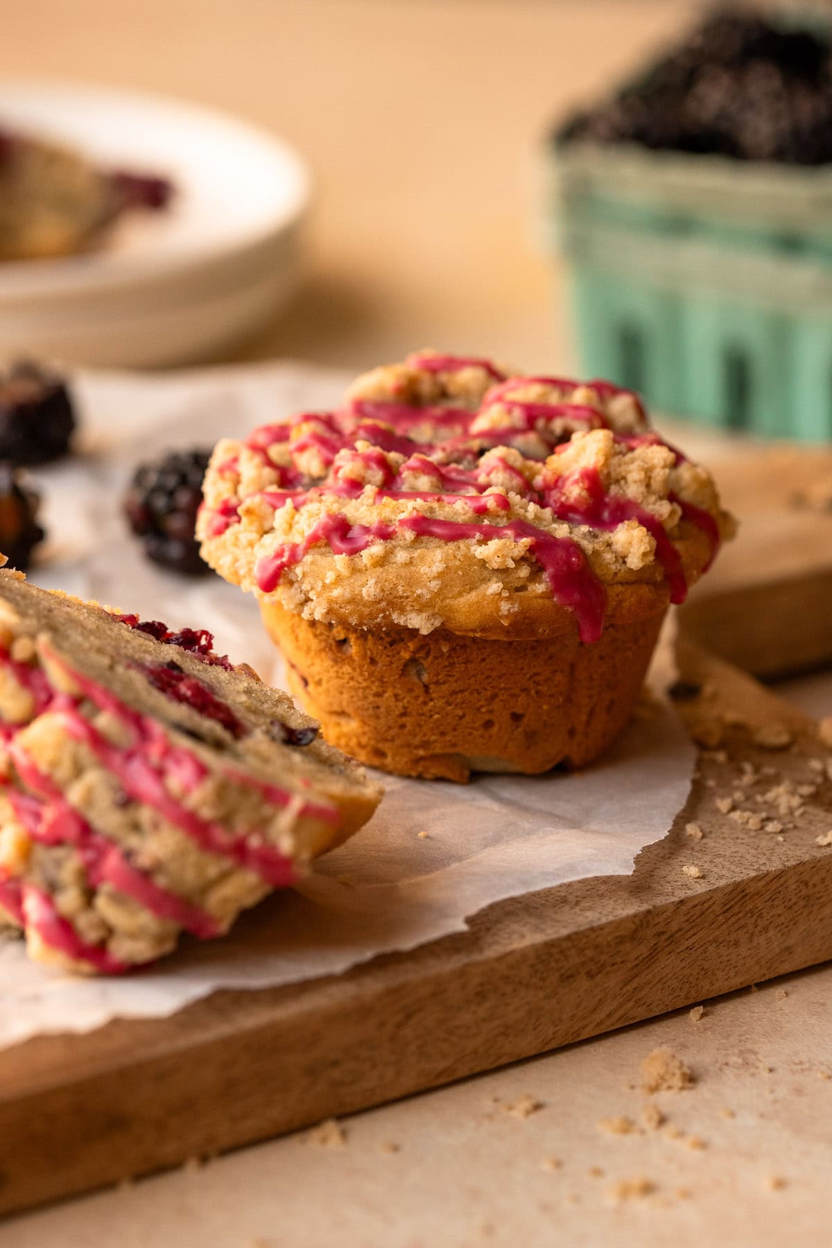 A muffin on a wooden board with another half muffin to the side and a basket of blackberries in the background.
