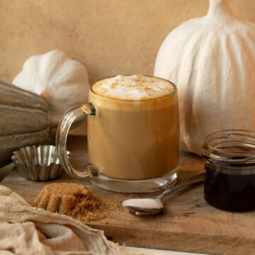 A latte in a clear mug garnished with brown sugar, with decorative pumpkins in the background.