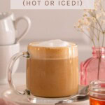 A latte served in a glass mug with a small jar of honey in the foreground. Text overlay reads "Easy Honey Latte Recipe (Served Hot or Iced!)