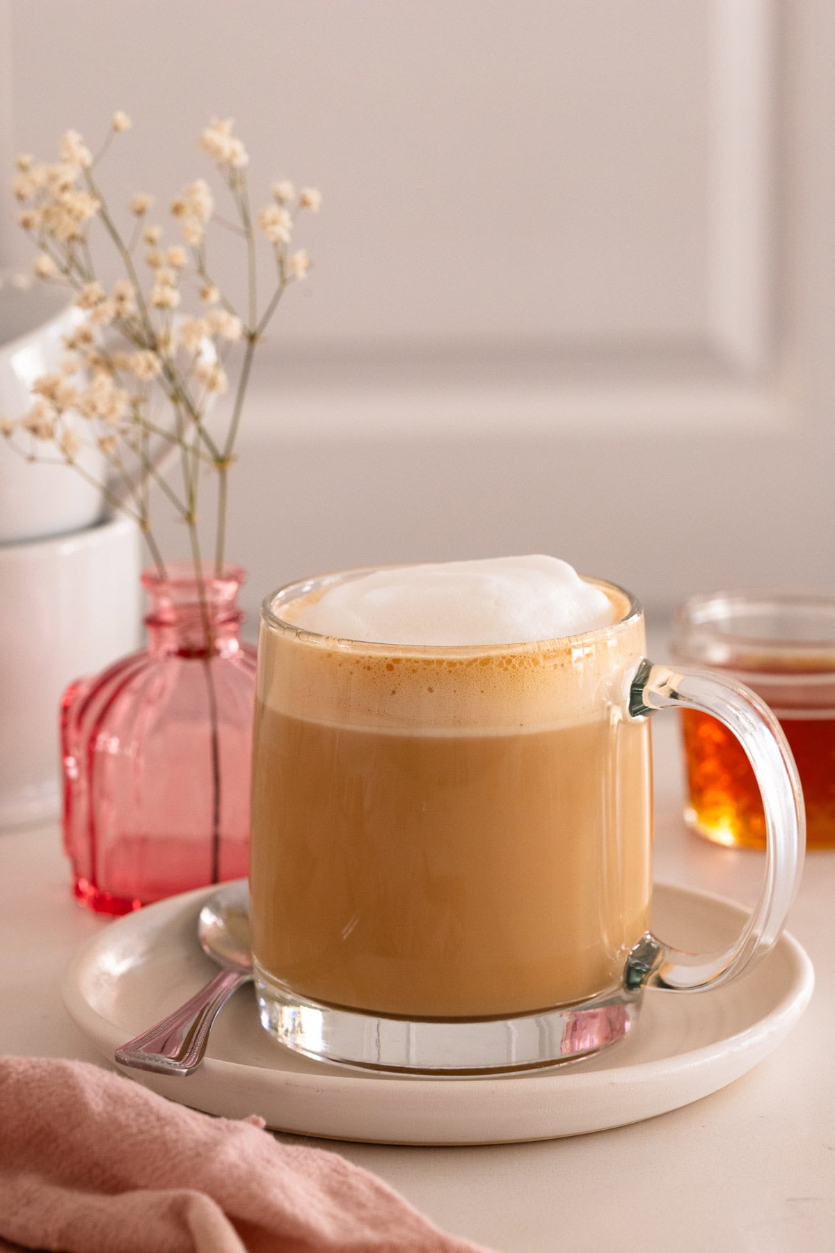 A latte in a glass mug on a white plate, with a bud vase and honey in the background.