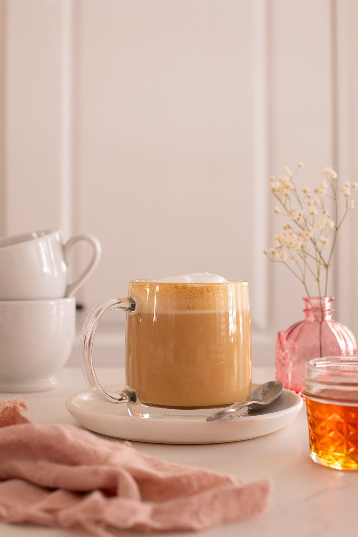 A latte in a glass mug on a small white plate, served with a spoon.