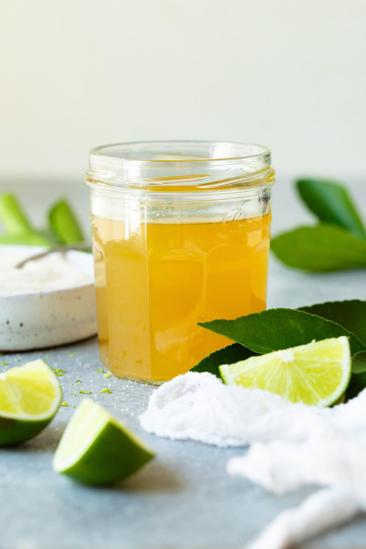 A jar filled with lime simple syrup, surrounded by lime wedges, lime tree leaves, and a bowl of sugar.