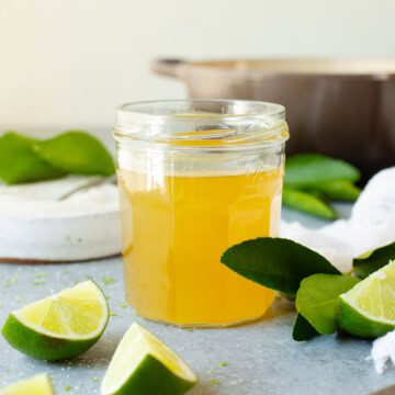 A jar of lime simple syrup alongside multiple lime wedges, with a pot used for cooking in the background.
