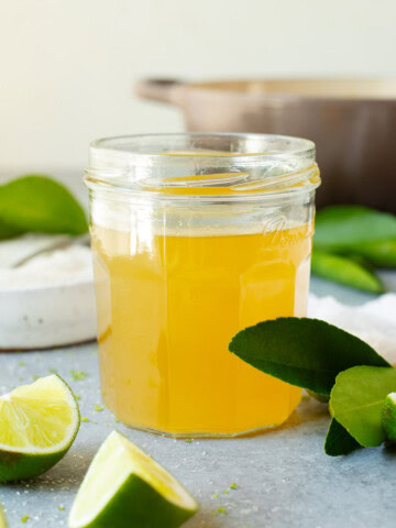 A jar of lime simple syrup alongside multiple lime wedges, with a pot used for cooking in the background.