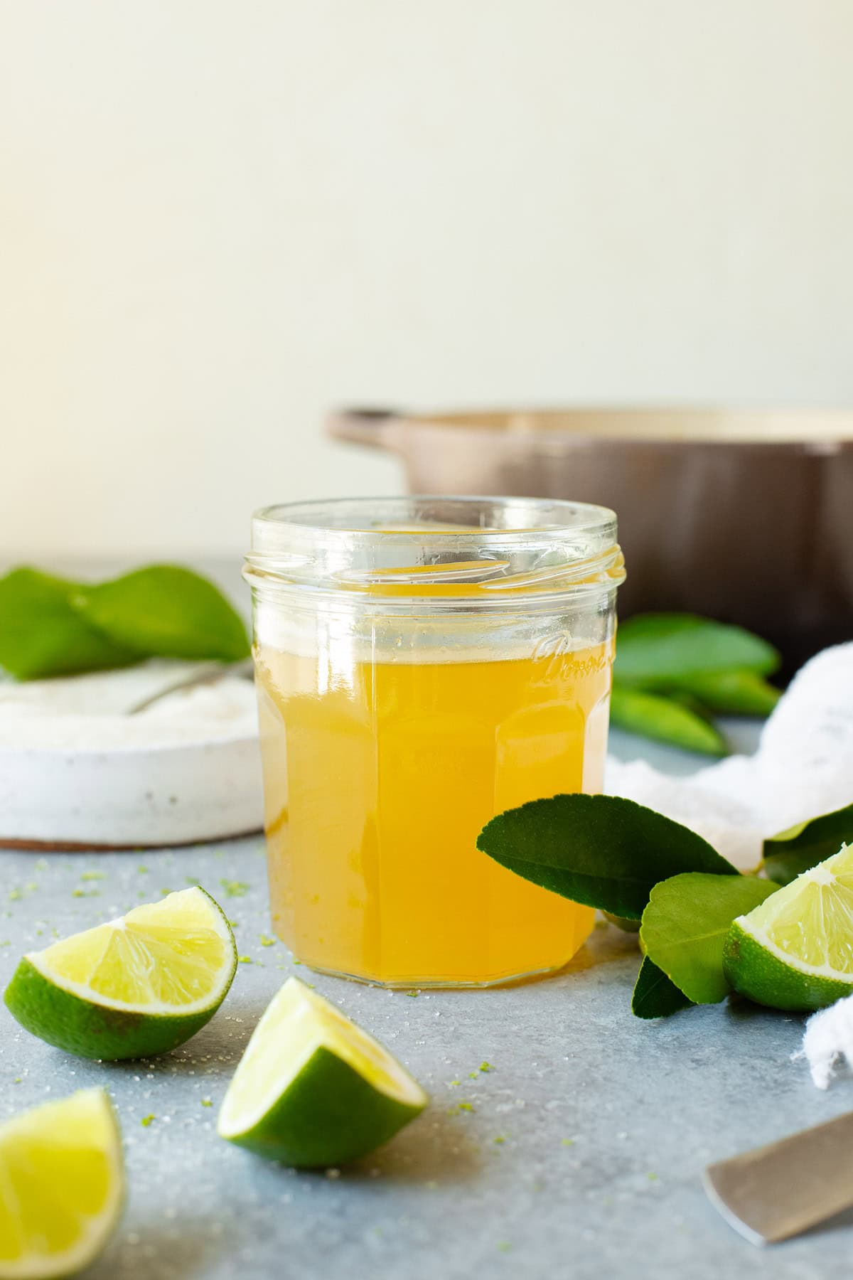 A jar of lime simple syrup alongside multiple lime wedges, with a pot used for cooking in the background.